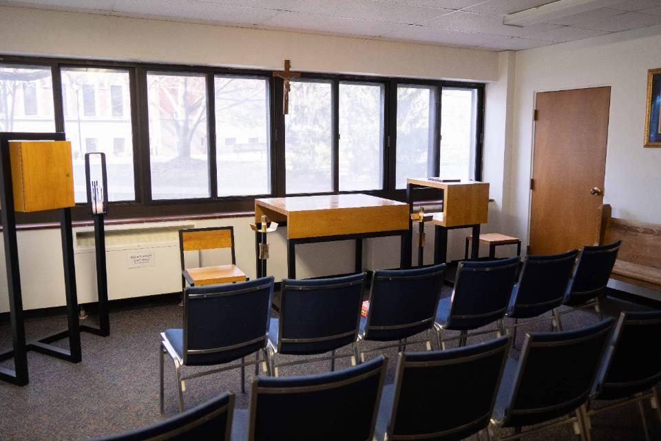 Chairs are seen from behind in two rows, facing a wooden altar table, chairs, crucifix and candle holder, along with a bank of windows.