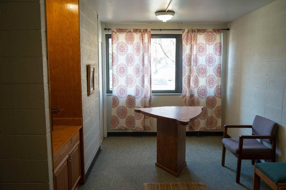 A room with a triangular table in the center, a few chairs and white curtains with a floral pattern.