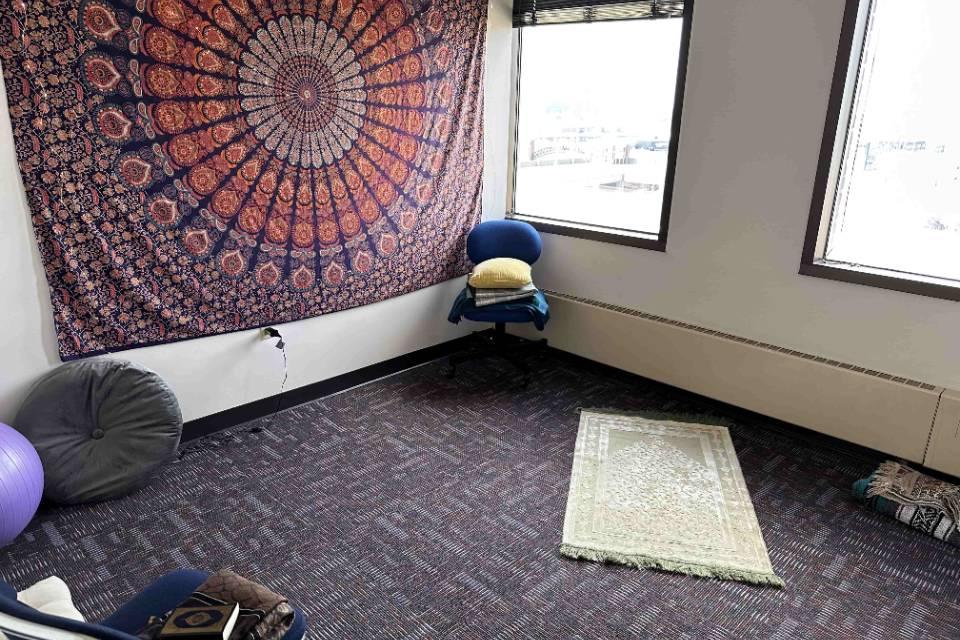 A room has a prayer rug on the floor as well as a mandala tapestry on one wall. Chairs and cushions are also visible.