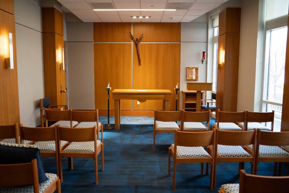 Chairs are seen in rows from behind with an altar at the front of the room. An abstract, modernist crucifix is on the wall.